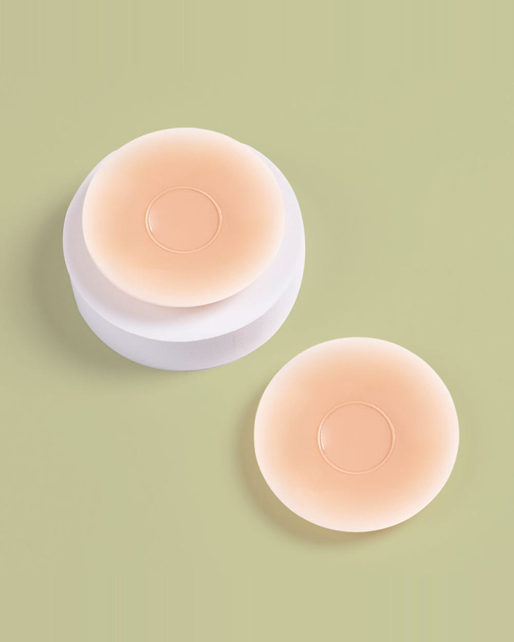 Two round Nudori Adhesive Nipple Covers on a sage background, one resting on its white storage case.