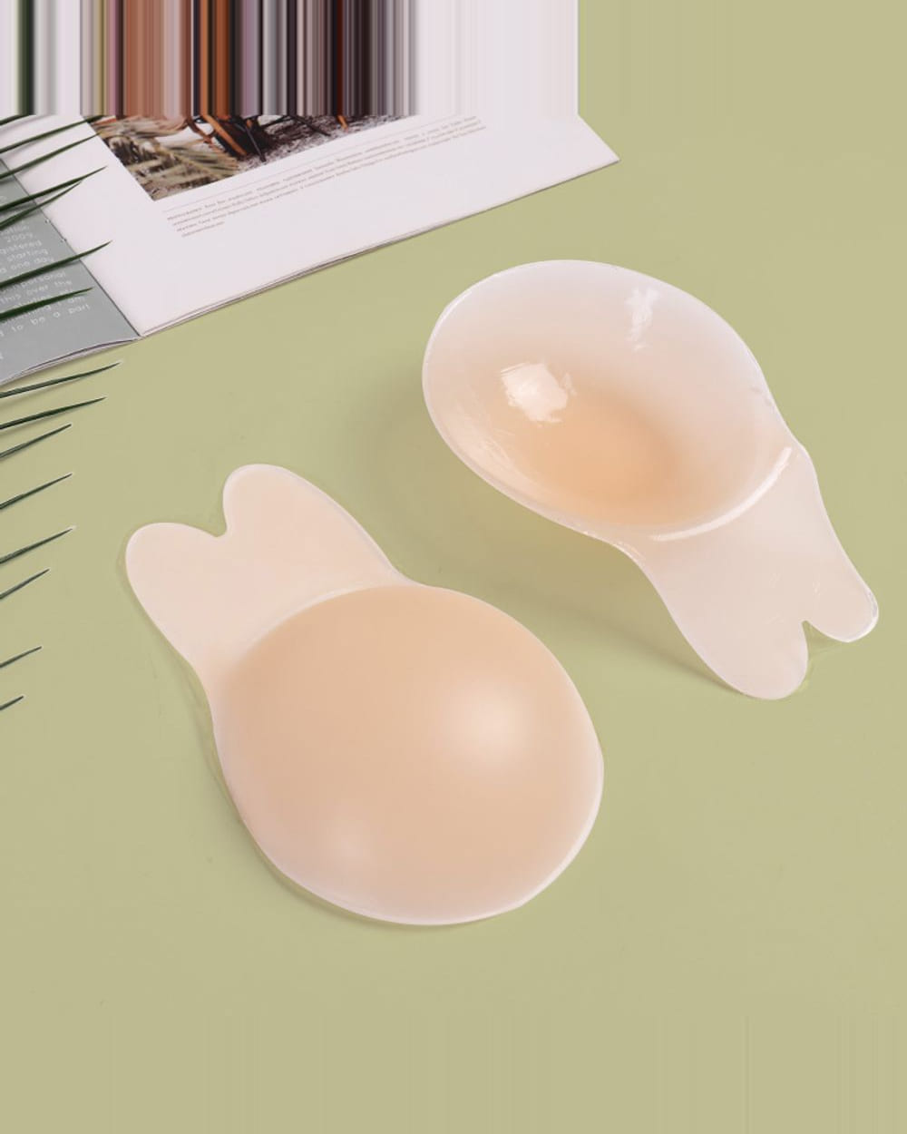 Two Nudori Bunny Lift-Up Nipple Covers on a sage background beside an open magazine, one lying face up and one turned to show its cupped underside.