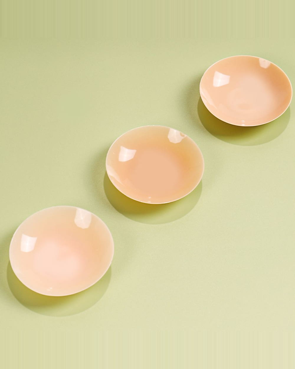 Three Nudori Non-Adhesive Nipple Covers arranged in a diagonal row on sage, their shallow domed shape catching the light.