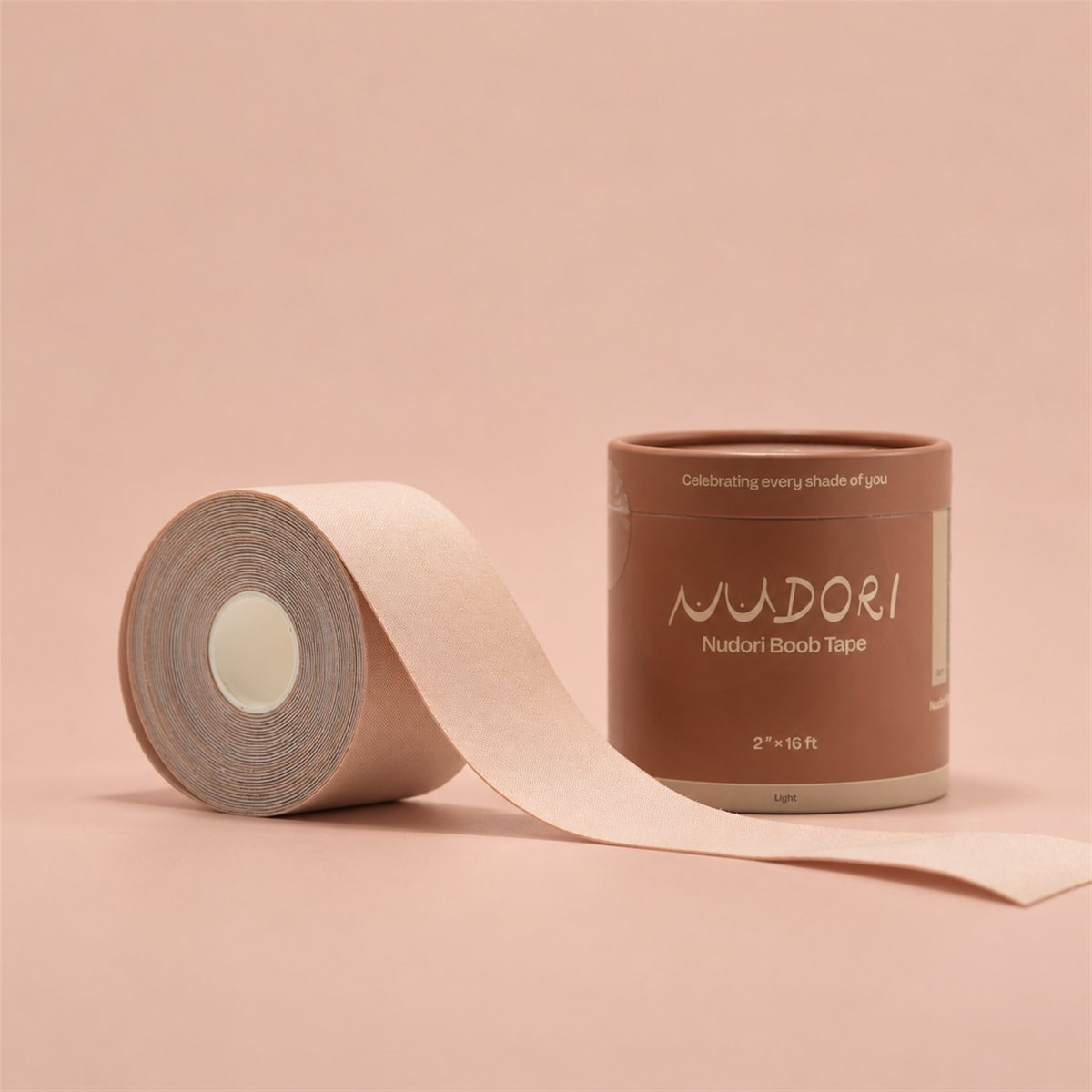Nudori Boob Tape roll in Light, partly unrolled beside its cylinder box on a nude studio background.